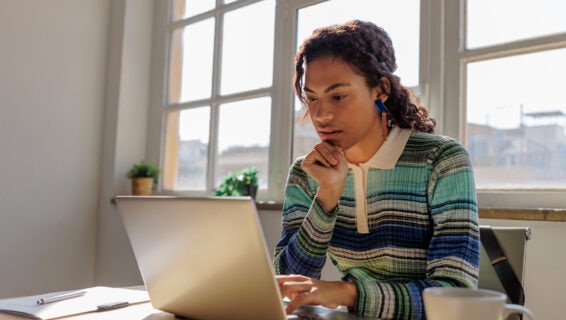 Woman sitting at a desk, focused on her laptop, with a notebook, pen, and coffee cup nearby, working in a bright office with large windows