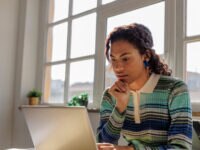 A young woman with braided hair, wearing a striped, long-sleeved polo shirt, sits at a desk working on a laptop. She rests her chin on her hand and looks intently at the screen. Sunlight streams through large windows behind her, with small potted plants visible on the windowsill.