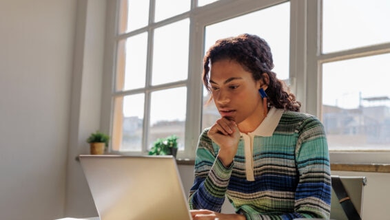 A young woman with braided hair, wearing a striped, long-sleeved polo shirt, sits at a desk working on a laptop. She rests her chin on her hand and looks intently at the screen. Sunlight streams through large windows behind her, with small potted plants visible on the windowsill.
