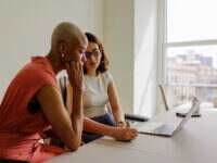 Two businesswomen in formal attire collaborating on a project, using a laptop and taking notes in a bright, modern office. One woman looks unsure.