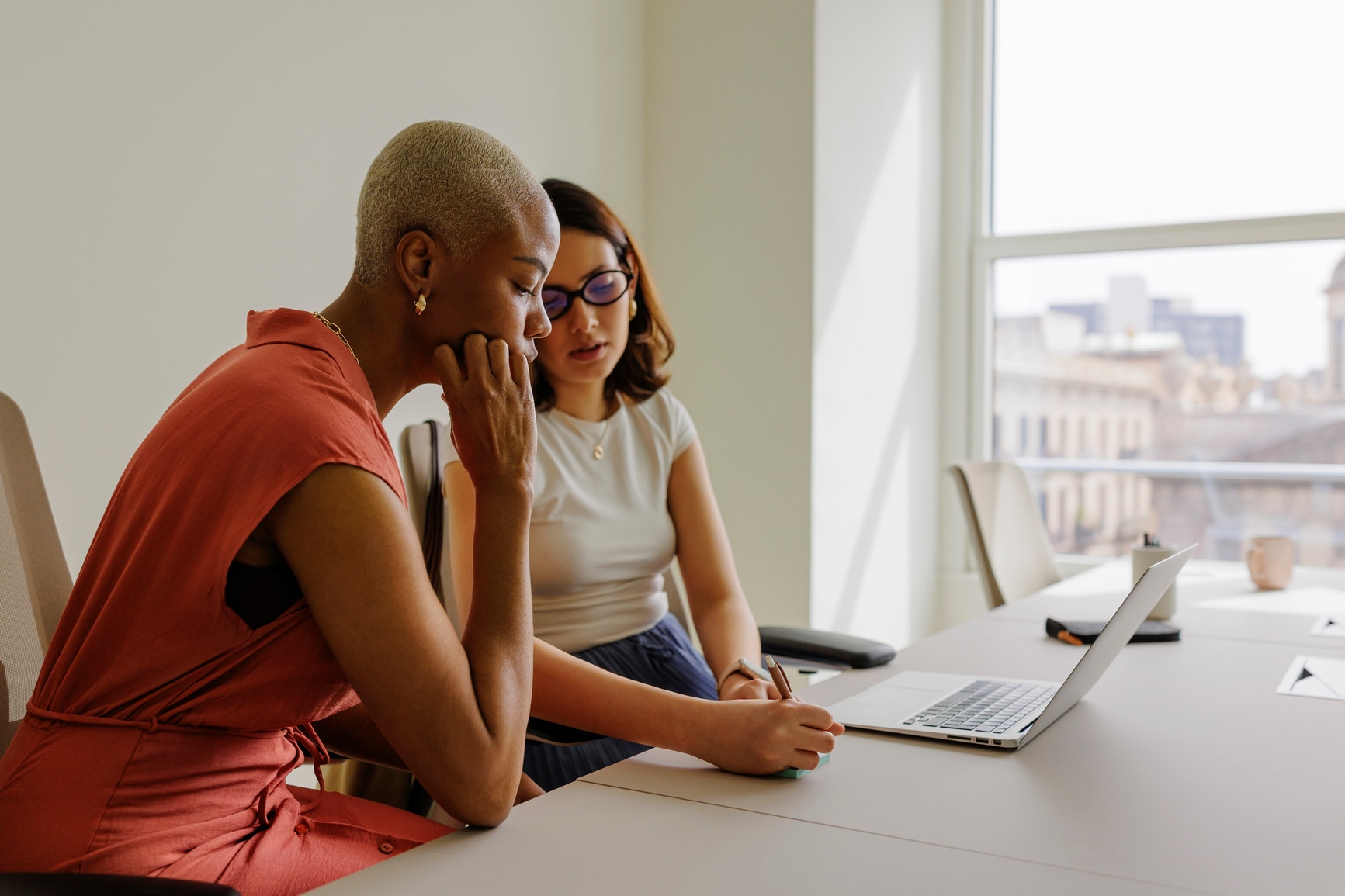 Two businesswomen in formal attire collaborating on a project, using a laptop and taking notes in a bright, modern office. One woman looks unsure.