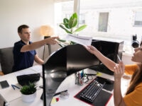 A man sitting at a desk in an office, reaching out to take a printed document from a woman seated across from him. The workspace features dual curved monitors, a keyboard, various stationery items, a small potted plant, and a large window letting in natural light. The woman holds a pen and appears to be explaining something.