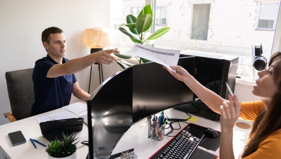 A man sitting at a desk in an office, reaching out to take a printed document from a woman seated across from him. The workspace features dual curved monitors, a keyboard, various stationery items, a small potted plant, and a large window letting in natural light. The woman holds a pen and appears to be explaining something.