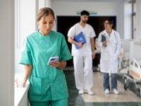 Female nurse using smartphone in hospital corridor while medical staff walks in the background