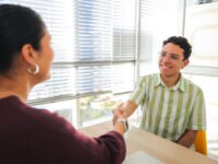 A young male job candidate smiles warmly while shaking hands with an HR agent, celebrating the end of a successful interview in a sunlit modern office. The bright natural light and casual atmosphere convey trust and openness