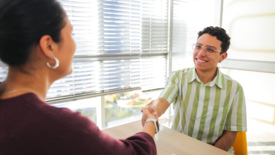 A young male job candidate smiles warmly while shaking hands with an HR agent, celebrating the end of a successful interview in a sunlit modern office. The bright natural light and casual atmosphere convey trust and openness