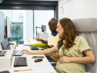 A pregnant woman sitting at a desk in a modern office, working on a laptop while a male colleague beside her reviews documents. Computer monitors, notebooks, and office supplies are visible on the desk.