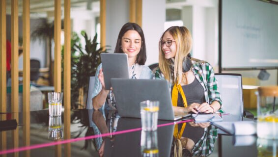 Two colleagues sitting at a desk, smiling while reviewing candidate profiles on a tablet and laptop, with glasses of water and notebooks in front of them.