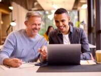 a younger and older colleague working together on a project, viewing something on the laptop screen