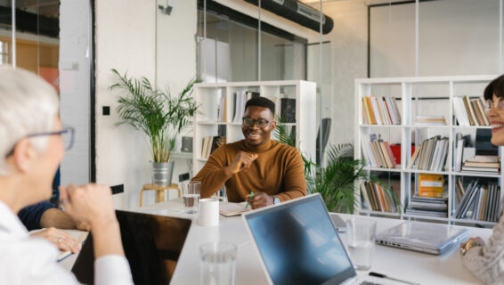 A smiling man in glasses and a brown jumper sits at a meeting table with colleagues, holding a pen and notebook. Laptops, glasses of water, and coffee mugs are on the table, with bookshelves and potted plants in the background of a modern office.