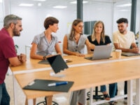 A group of five colleagues gathered around a high desk in a modern office, engaged in discussion. Two women are seated, one of whom is pointing at a laptop screen, while the others stand or lean in attentively. Notebooks, a tablet, and a takeaway coffee cup are on the desk. The office space features glass walls and a bright, open layout.