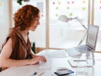 A woman with curly red hair working at a desk in a bright, modern office. She is typing on a wireless keyboard, with a laptop placed on a stand in front of her. The desk also holds a mouse, tablet, notebook, and a glass of water. Colourful sticky notes are visible on the glass wall in the background.