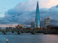Aerial view of the London skyline at dusk, with the Shard rising above the Thames and Southwark Bridge, under a dramatic cloudy sky.