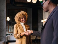 Two professionals shaking hands in a stylish, modern setting. The woman, smiling warmly, is wearing a tan blazer and white turtleneck, while the man, slightly out of focus, is dressed in a dark suit. The background features a softly lit dining area with elegant decor and wall-mounted lights.