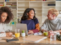 A bright, modern workspace with three young colleagues seated at a wooden table: on the left, a woman with voluminous curly hair writes in a notebook; centre, a woman with dark hair smiles as she types on a laptop; and on the right, a fair-haired man in a chunky knit jumper jots notes. The table is strewn with glasses of orange and green juice, a small potted cactus, headphones, an apple and various papers, set against a backdrop of white shelving filled with books and decor.