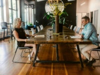 Work partners seated in a huge wooden table at work surrounded by notepads and notebooks