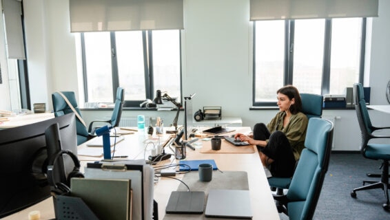 A woman with earphones uses a computer sitting at her desk in the office alone. Concentrated look. Daylight.