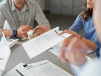 Close-up of a business meeting, with one person holding and pointing at a printed document clipped to a folder. Other participants are seated around the table, with laptops, pens, and takeaway coffee cups visible. The focus is on the discussion of the document, with hands and paperwork in the foreground.