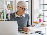 Senior female employee preparing herself for a meeting She is sitting at a desk, writing some notes in her notebook, her laptop and smartphone next to her.