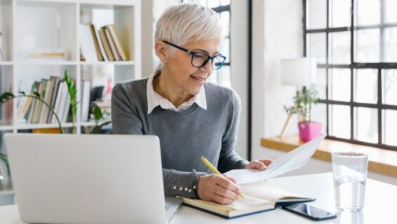 Senior female employee preparing herself for a meeting She is sitting at a desk, writing some notes in her notebook, her laptop and smartphone next to her.