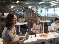 An image of a woman holding a laptop in a modern office, with colleagues eating pizza in the background.