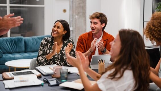Group of diverse colleagues sitting at table and applauding excited with great news and business achievements while having meeting in modern coworking space.