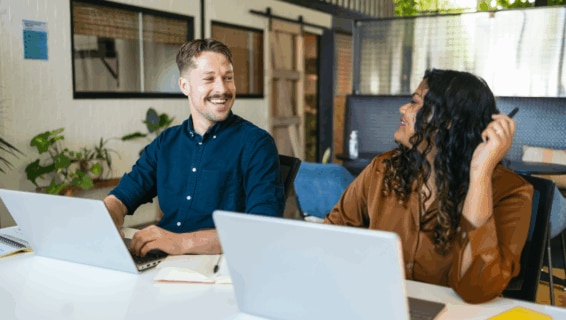 Two cheerful coworkers talking and sharing ideas in the office while working with laptops, highlighting the value of an engaged, motivated, and loyal workforce