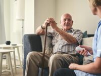 An image of an elderly man holding a walking stick, alongside a nurse who is caring for him.