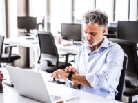 A man in an office surrounded by empty desks, checking his watch.