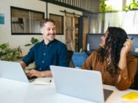 Two cheerful coworkers talking and sharing ideas in the office while working with laptop, highlighting the importance of collaboration, communication, and employee satisfaction to long term retention.