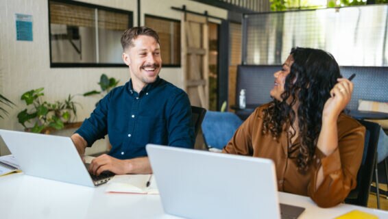 Two cheerful coworkers talking and sharing ideas in the office while working with laptop, highlighting the importance of collaboration, communication, and employee satisfaction to long term retention.